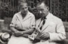 Eine Nahaufnahme von Katia und Thomas Mann. Thomas, rechts im Bild, mit runder Brille, ist auf ein geöffnetes Buch in seinen Händen konzentriert und scheint darüber zu sprechen. Katia, links im Bild, schaut ebenfalls in das Buch und folgt aufmerksam. Beide