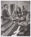 Thomas Mann aufgestützt auf das Piano im Gespräch mit dem Pianisten Bruno Walter, im Hintergrund Katia Mann mit Lotte Walter und Klaus Mann.