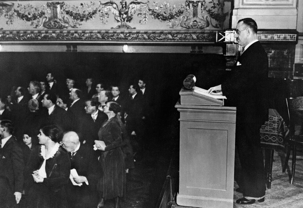 ALT-Text: Schwarz-Weiß-Fotografie von Thomas Mann während seiner Rede „Deutsche Ansprache. Ein Appell an die Vernunft" am 17. Oktober 1930 im Beethoven-Saal in Berlin. Mann steht am linken Bildrand am Rednerpult und blickt in das Publikum, das sich von ihm abwendet und nach hinten in den Saal schaut. Die Aufnahme zeigt die angespannte Atmosphäre während der Rede, bei der es zu nationalsozialistischen Störungen kam.